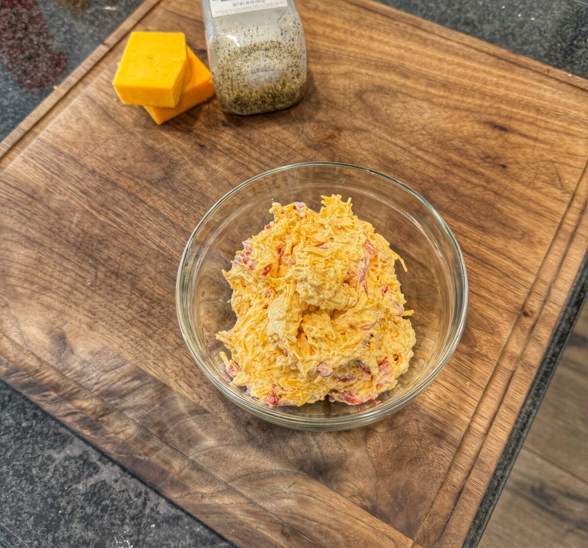 Homemade pimento cheese placed in a clear glass bowl on a wooden cutting board with blocks of cheddar cheese nearby.