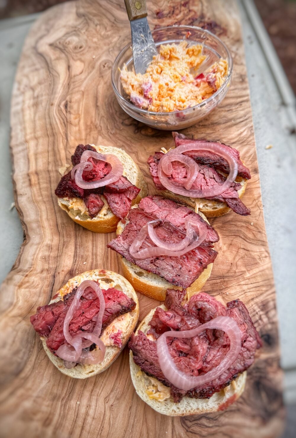 Toasted crostini topped with sliced steak, pimento cheese, and pickled red onions on a wooden serving board.