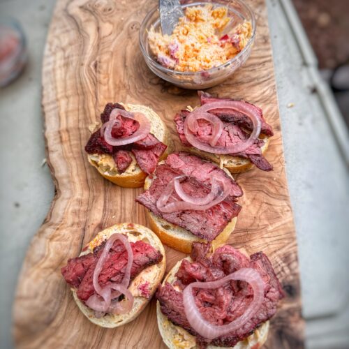Toasted crostini topped with sliced steak, pimento cheese, and pickled red onions on a wooden serving board.