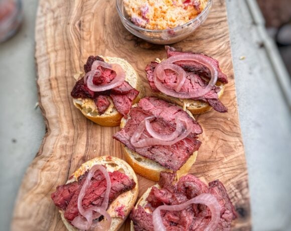 Toasted crostini topped with sliced steak, pimento cheese, and pickled red onions on a wooden serving board.