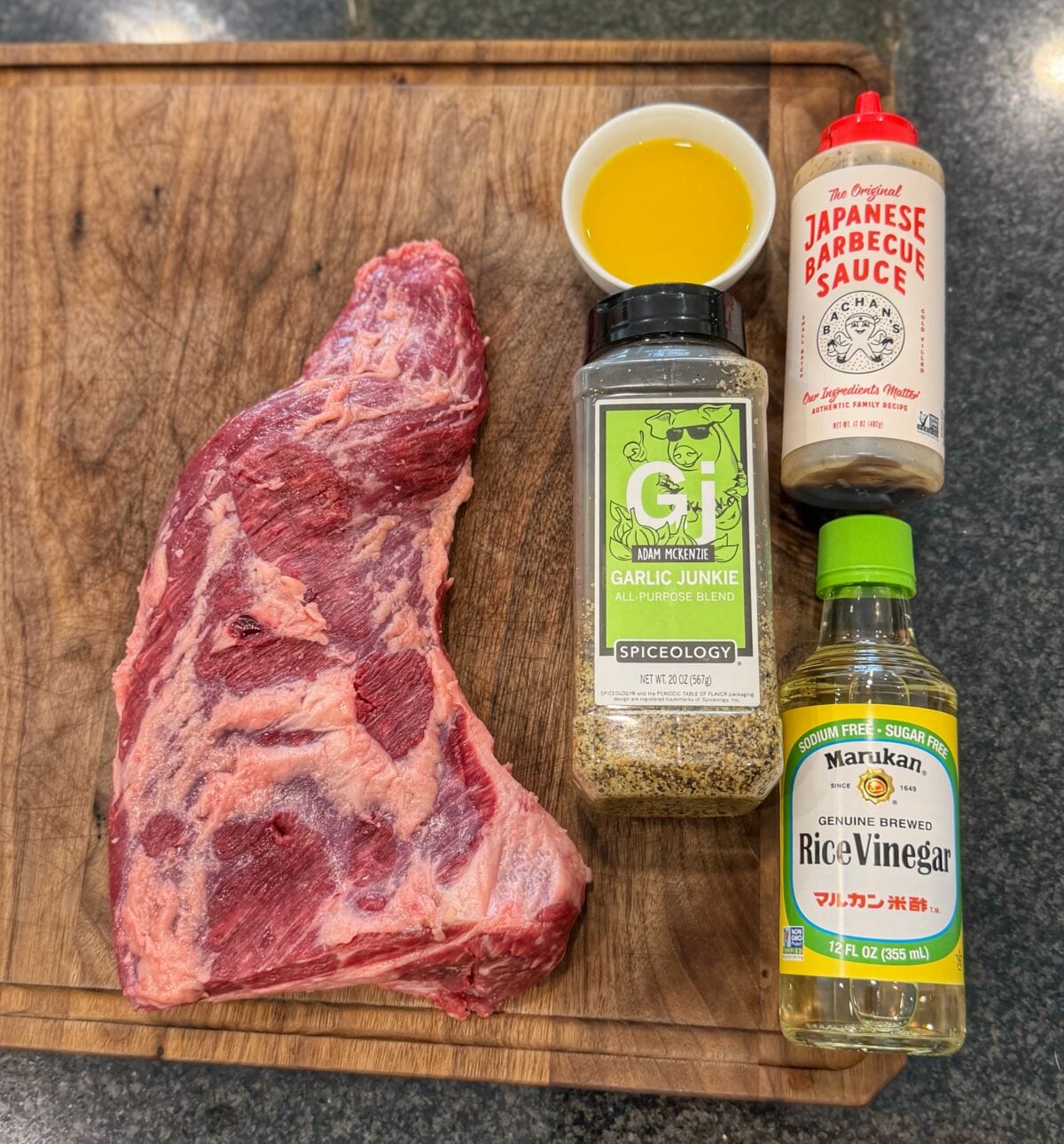 Tri tip roast on a cutting board surrounded by orange juice, Bachan’s Japanese BBQ Sauce, rice vinegar, Garlic Junkie seasoning, and Pit Boss Classic Steakhouse Rub.