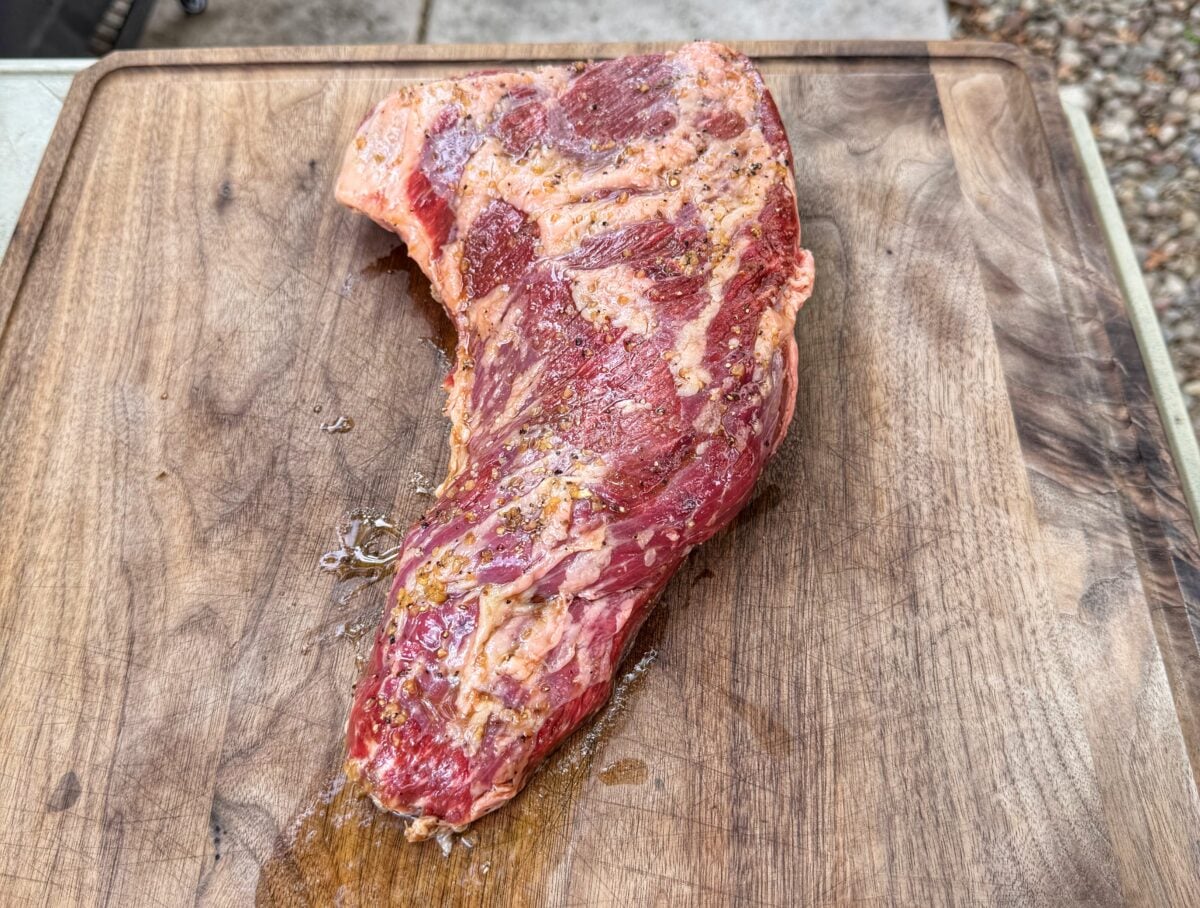 Raw tri tip roast coated in marinade resting on a wooden cutting board.