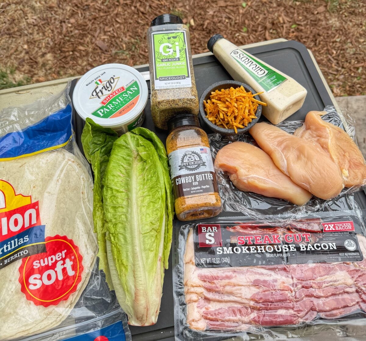Ingredients for grilled chicken Caesar wraps including tortillas, romaine, chicken, bacon, parmesan, Caesar dressing, seasoning, and jalapeño potato straws.
