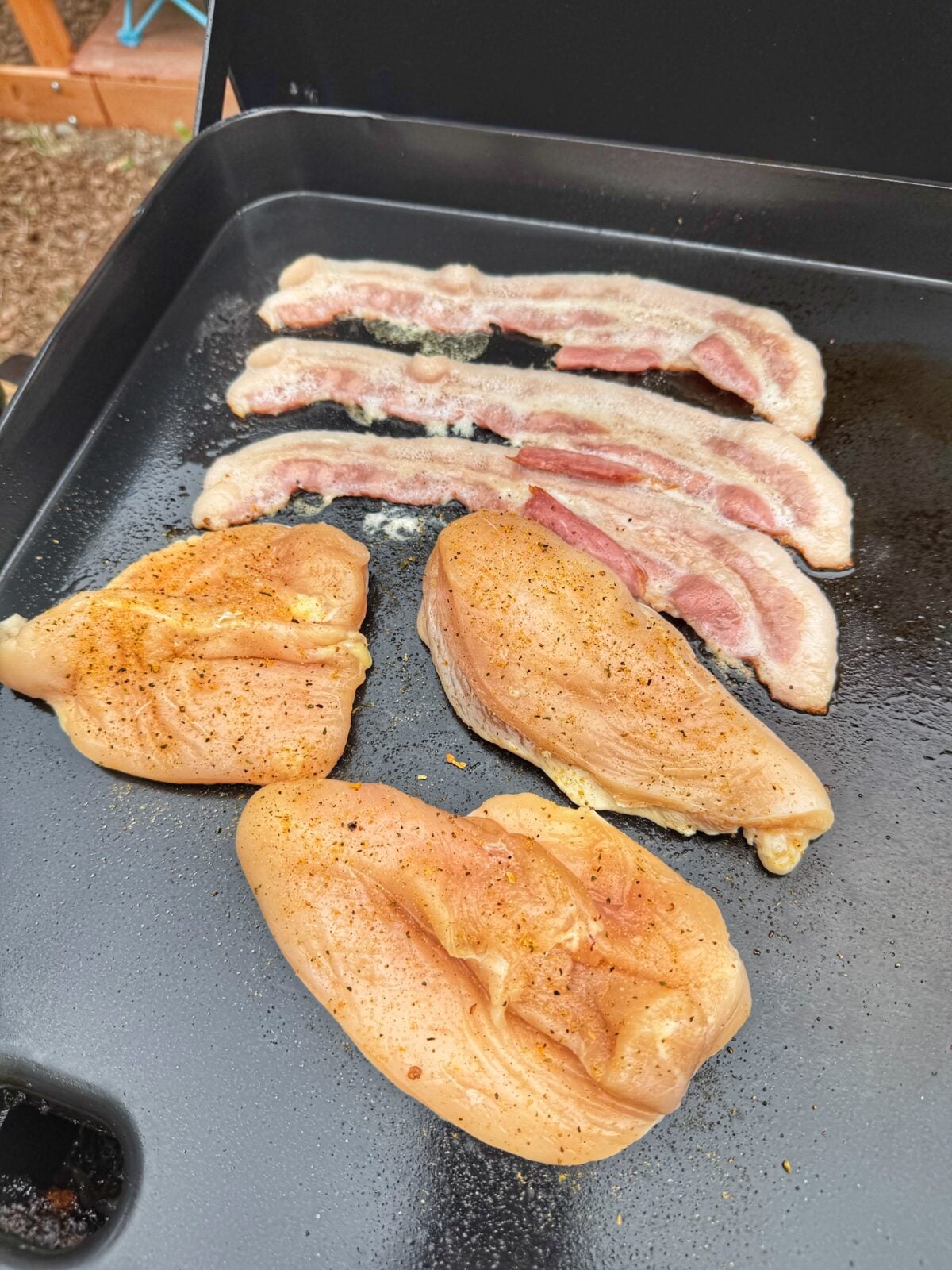 Butterflied chicken breasts and steak-cut bacon cooking on a flat top griddle.