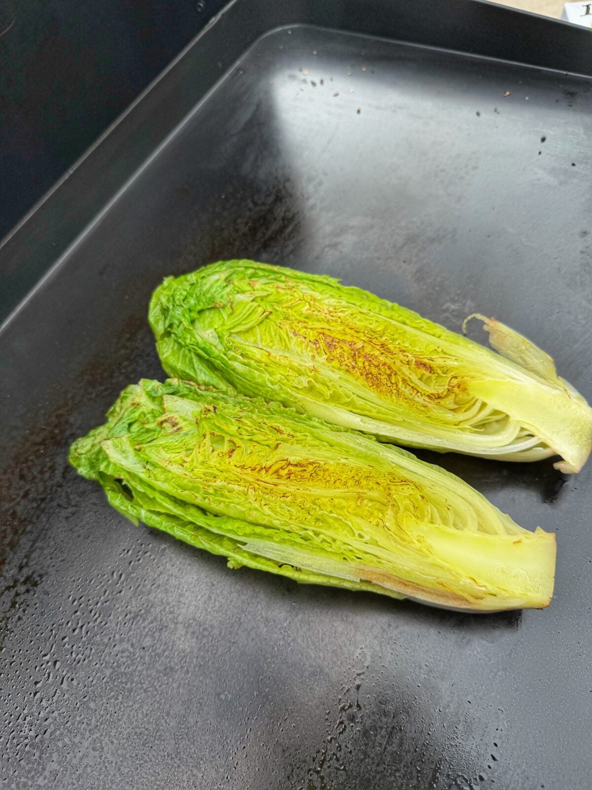 Halved romaine hearts grilling cut side down on a flat top griddle.