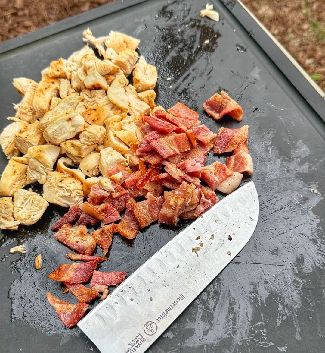 Cooked chicken breast and chopped bacon on a cutting board with a chef knife.