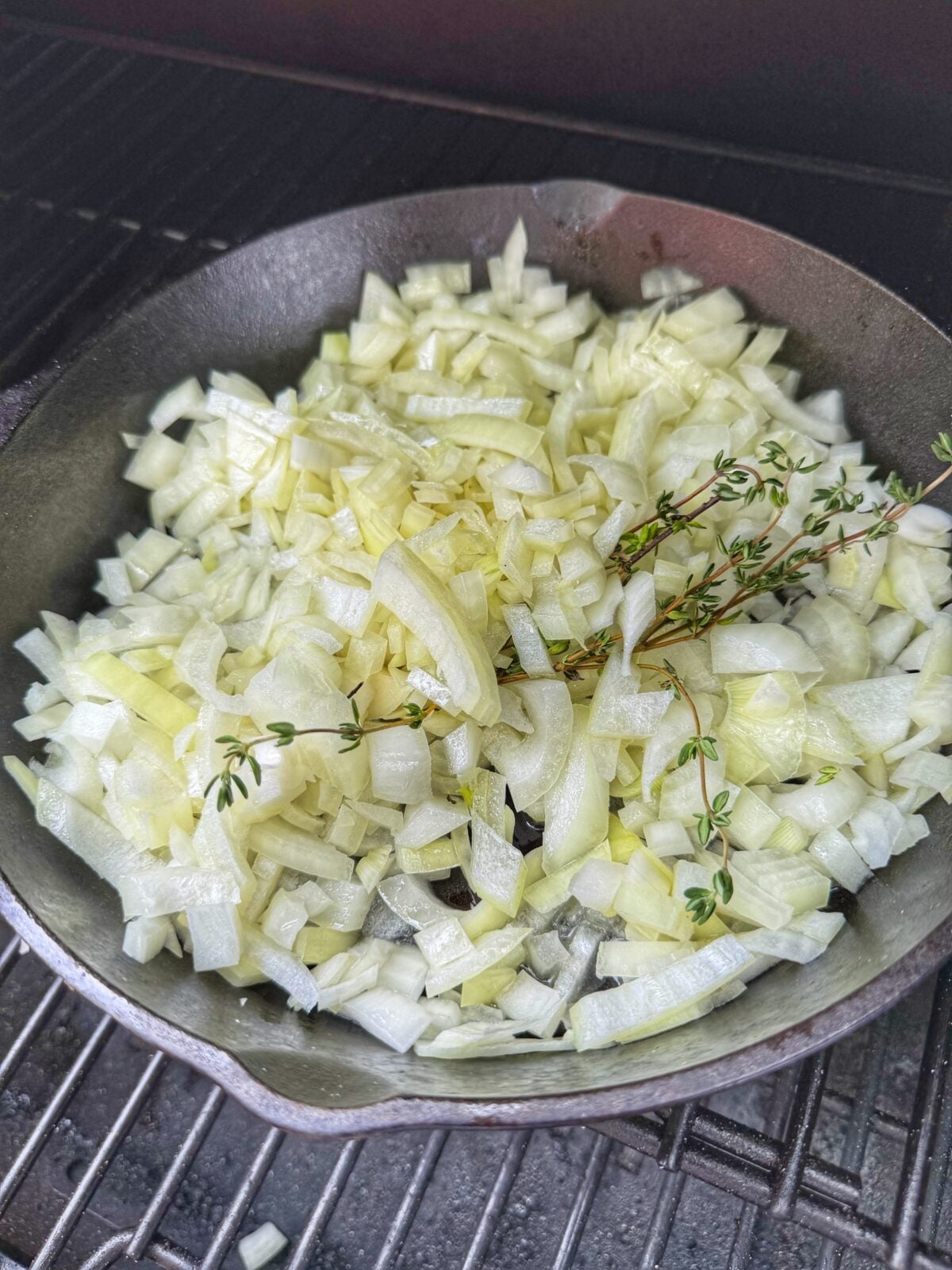 Diced yellow onions and fresh thyme cooking in a cast iron skillet on a pellet grill.