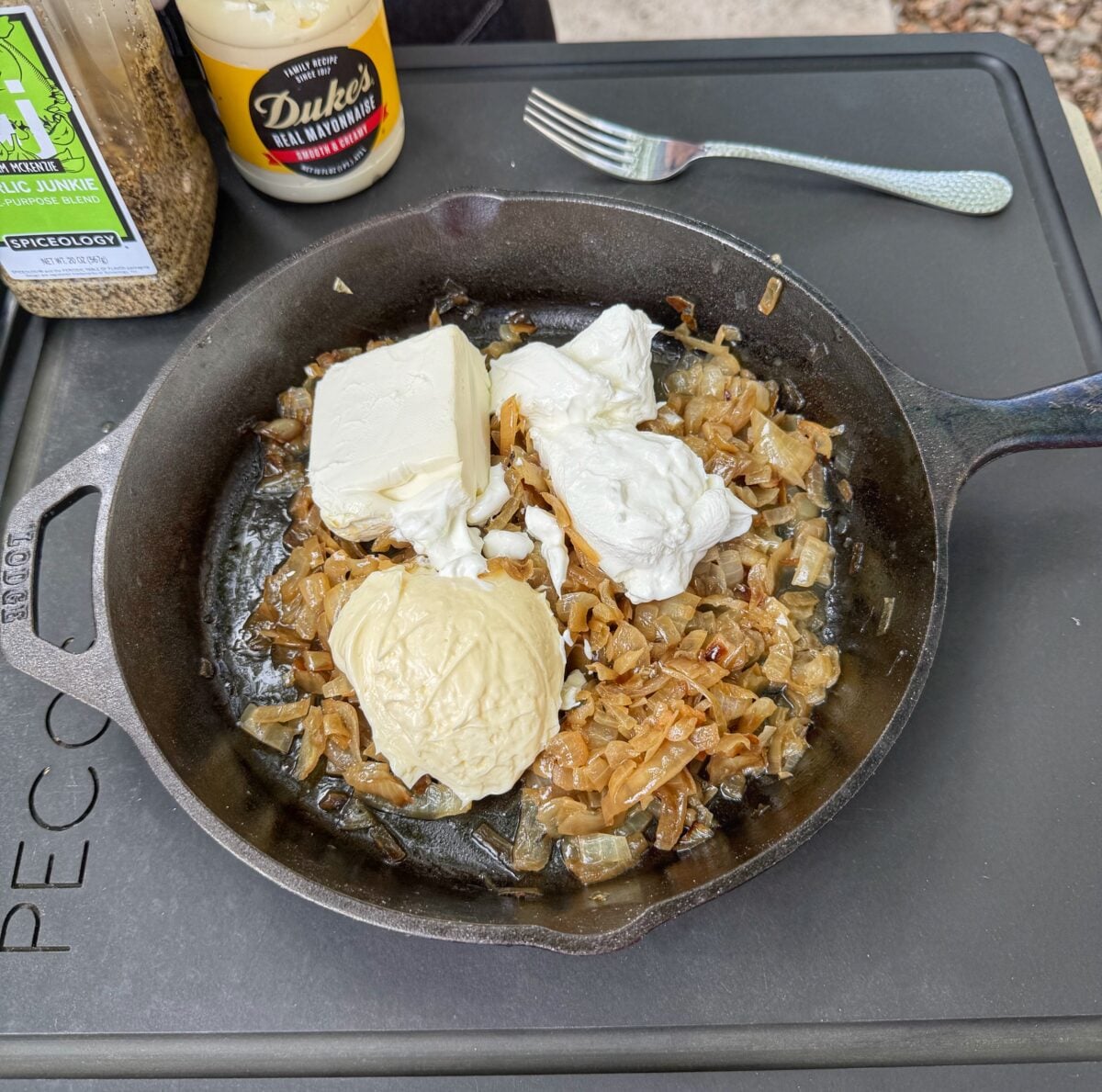 French onion dip mixture being stirred in a cast iron skillet with Duke’s mayonnaise and Garlic Junkie seasoning nearby.