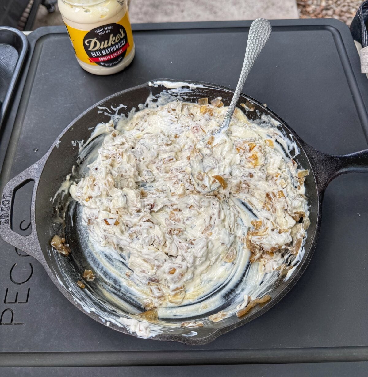 Caramelized onions in a cast iron skillet topped with cream cheese, sour cream, and mayonnaise beside Garlic Junkie seasoning and Duke’s mayo.