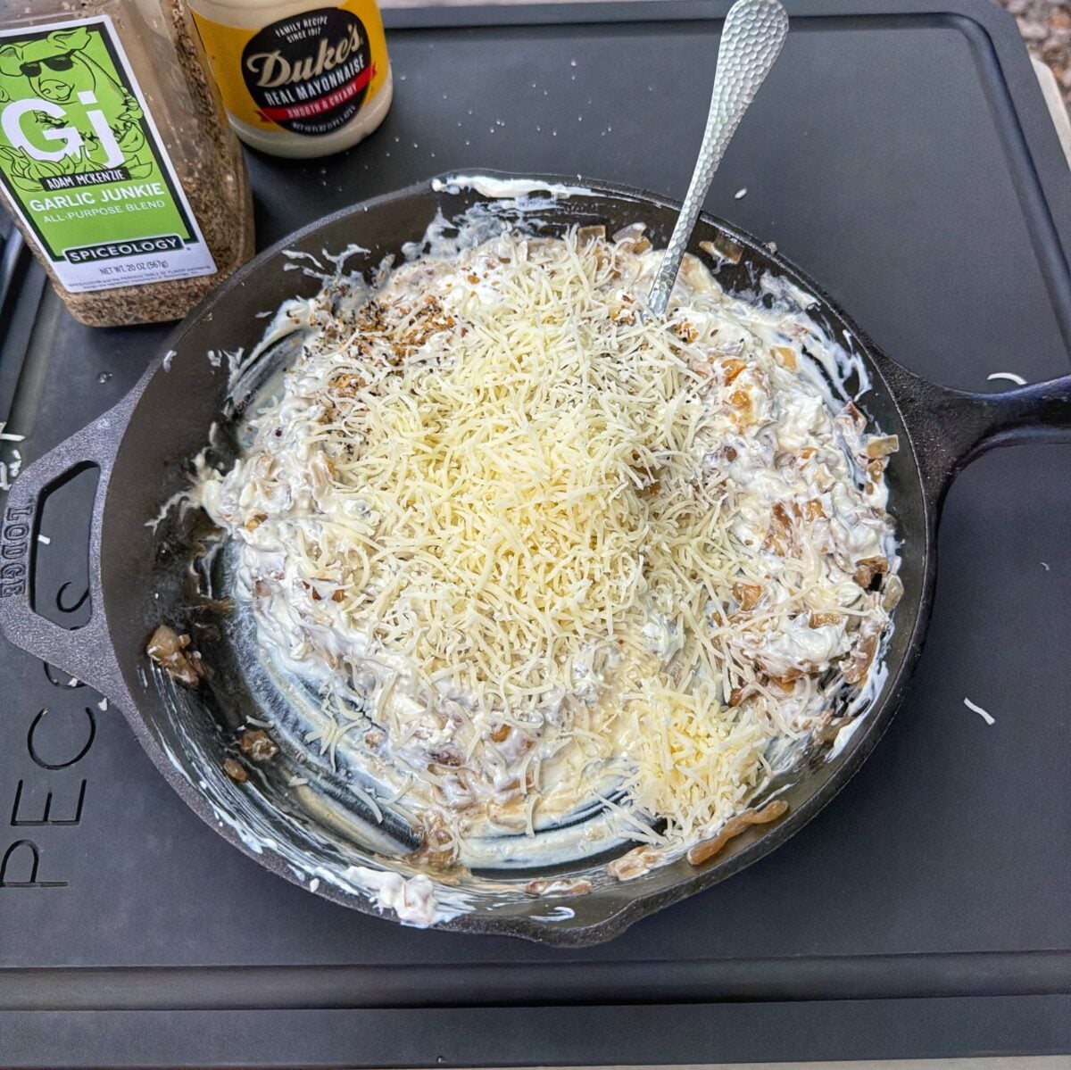French onion dip mixture in a cast iron skillet topped with shredded Swiss cheese and Garlic Junkie seasoning.