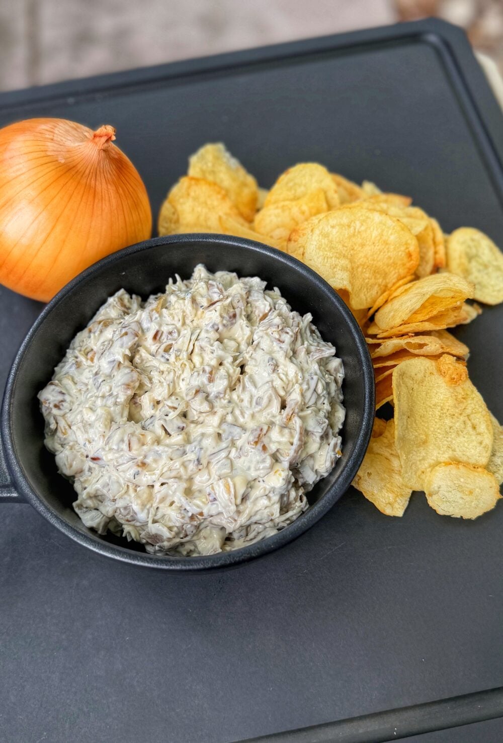 Homemade French onion dip in a black bowl served with potato chips and a whole yellow onion on a black tray.