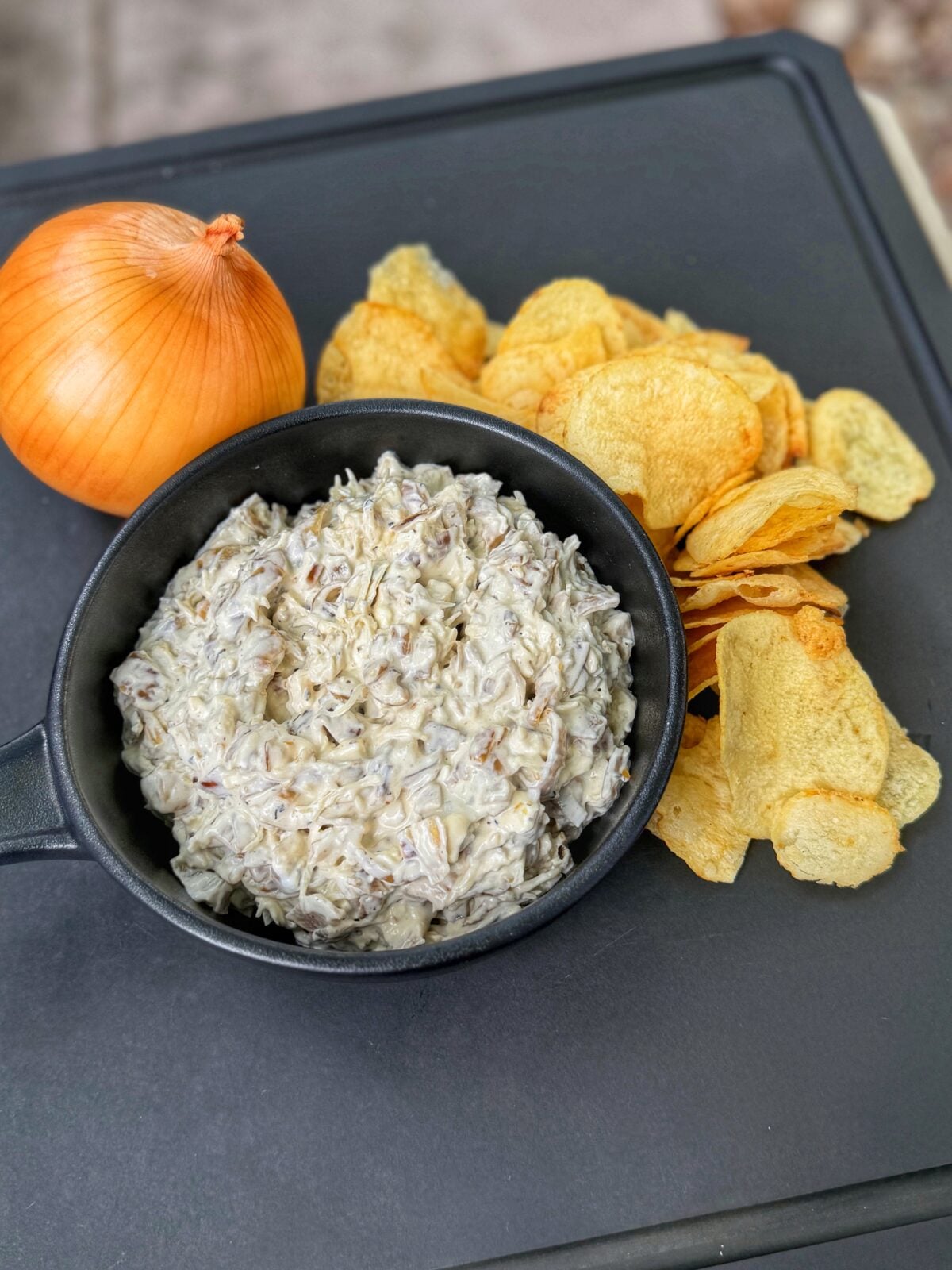 Homemade French onion dip in a black bowl served with potato chips and a whole yellow onion on a black tray.