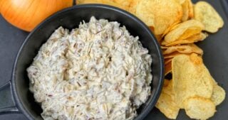 Homemade French onion dip in a black bowl served with potato chips and a whole yellow onion on a black tray.
