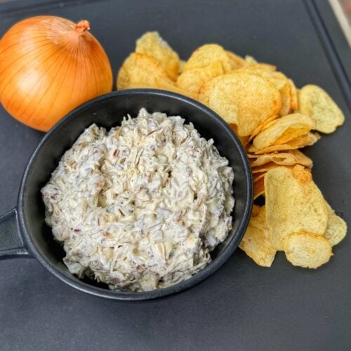 Homemade French onion dip in a black bowl served with potato chips and a whole yellow onion on a black tray.