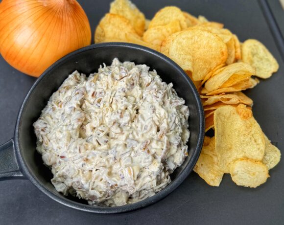 Homemade French onion dip in a black bowl served with potato chips and a whole yellow onion on a black tray.