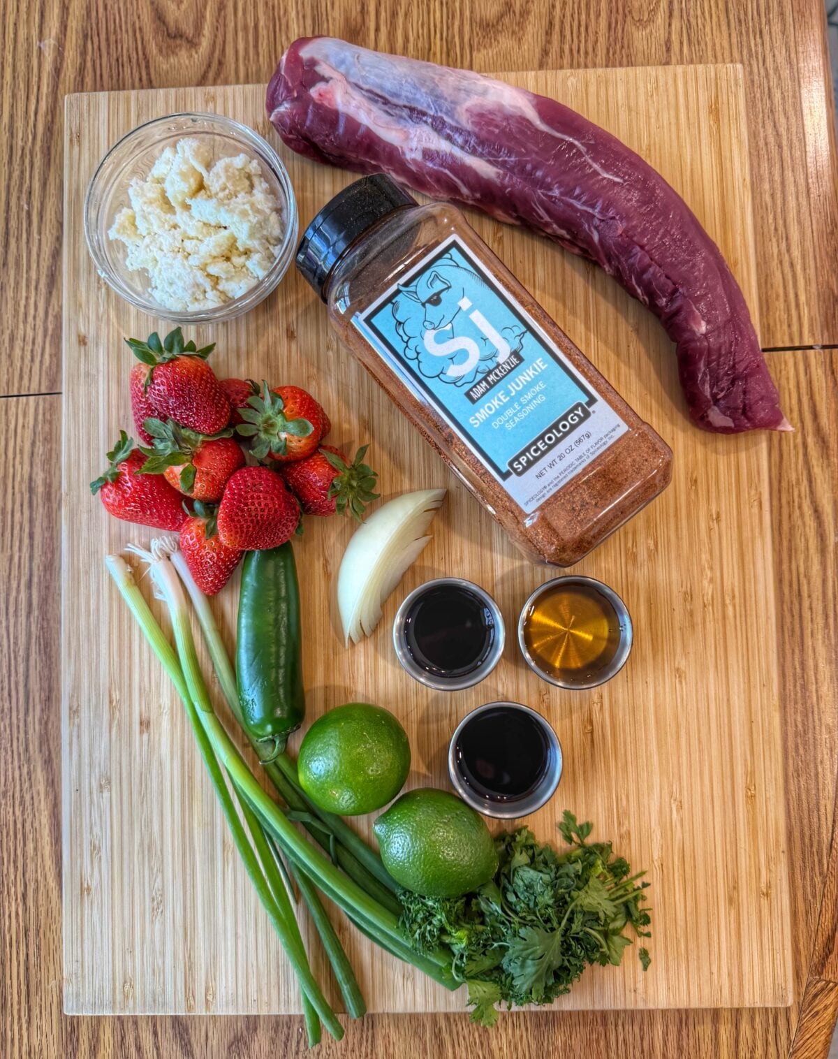 Pork tenderloin, Smoke Junkie seasoning, strawberries, cotija cheese, jalapeño, cilantro, limes, onion, and sauce ingredients arranged on a wooden board.