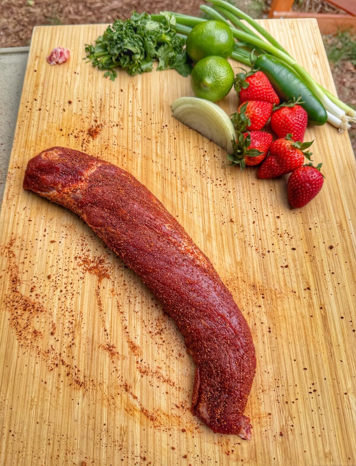 Raw pork tenderloin coated in Smoke Junkie seasoning on a cutting board beside strawberries, jalapeño, cilantro, limes, onion, and green onions.