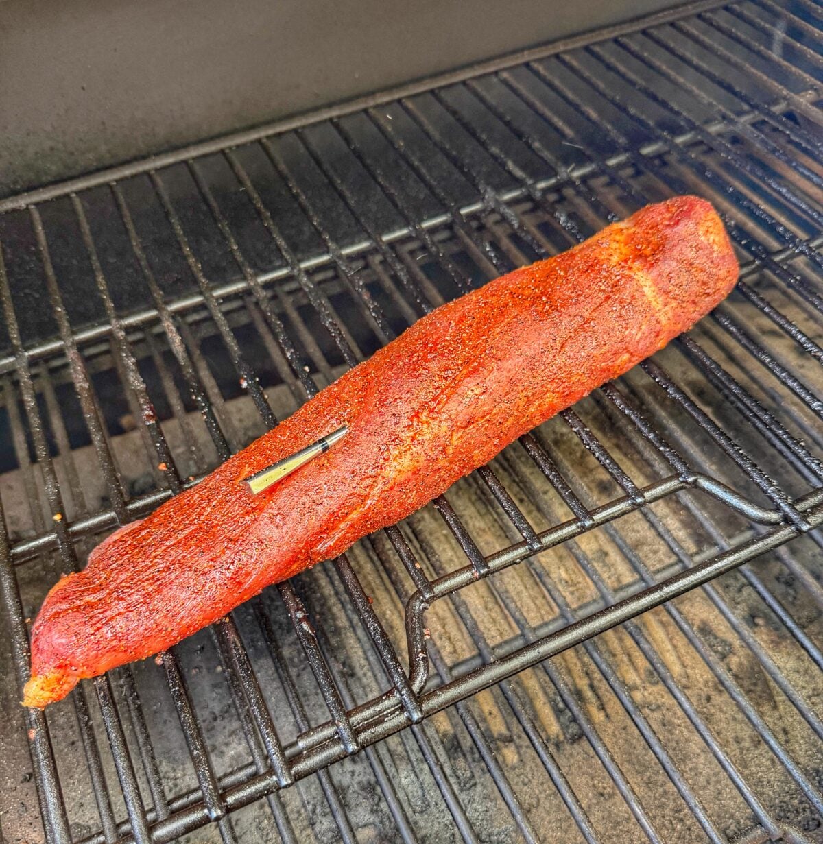 Seasoned pork tenderloin smoking on the grates of a Pit Boss Navigator pellet grill with a meat thermometer inserted.