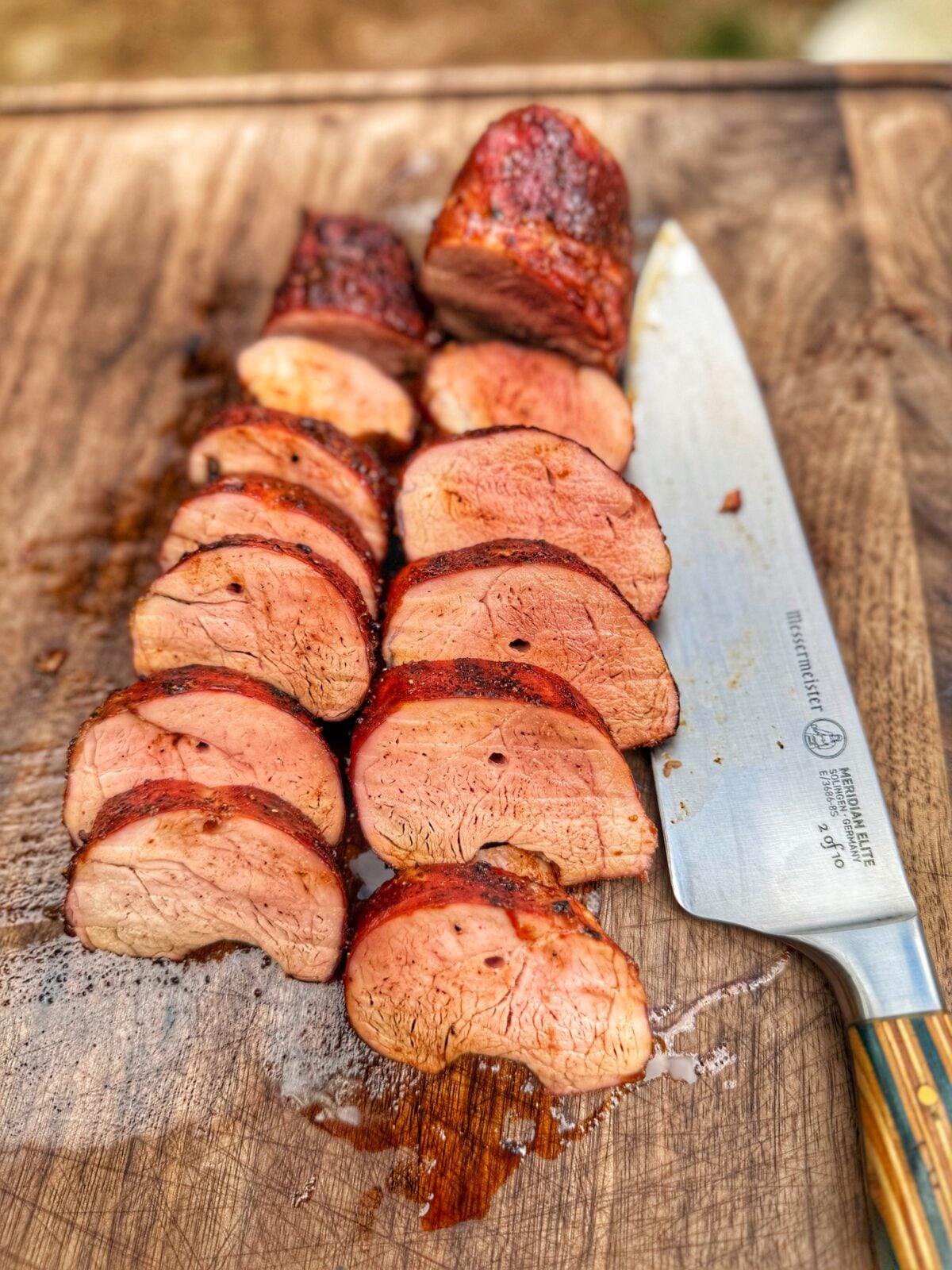 Sliced smoked pork tenderloin on a wooden cutting board next to a chef knife with juices pooling beneath the meat.