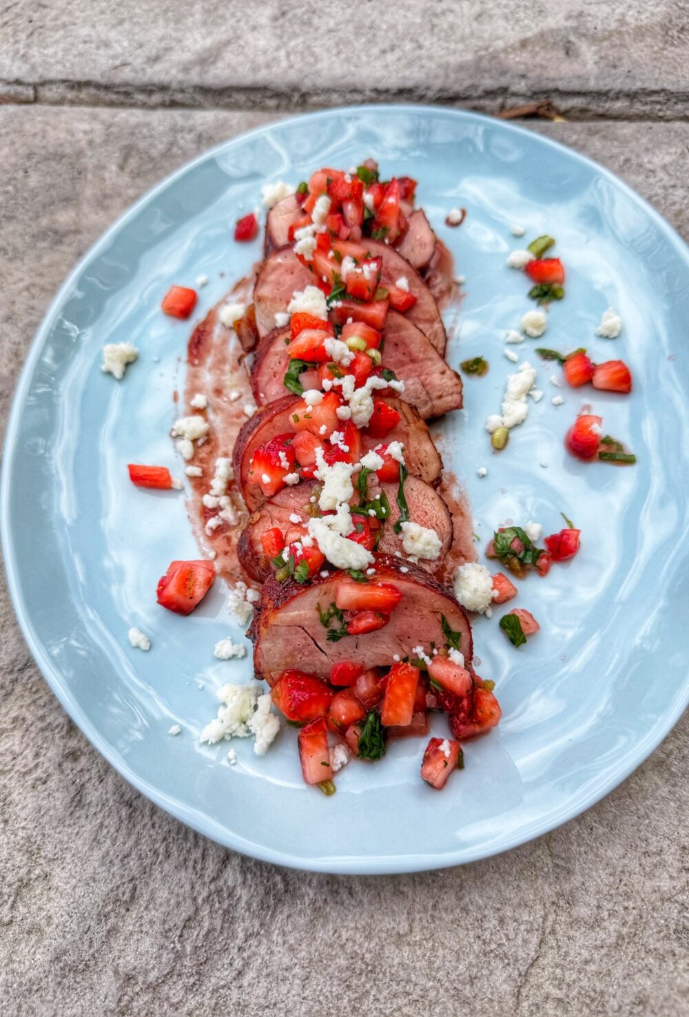 Sliced pork tenderloin topped with strawberry salsa and crumbled cotija cheese served on a light blue plate.