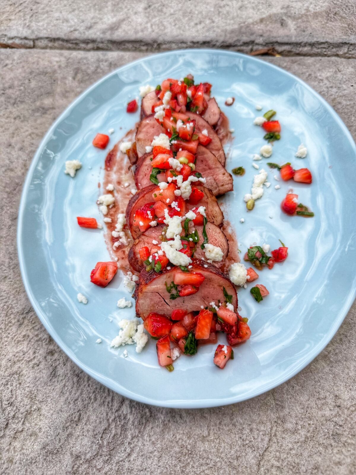 Sliced pork tenderloin topped with strawberry salsa and crumbled cotija cheese served on a light blue plate.