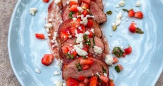 Sliced pork tenderloin topped with strawberry salsa and crumbled cotija cheese served on a light blue plate.