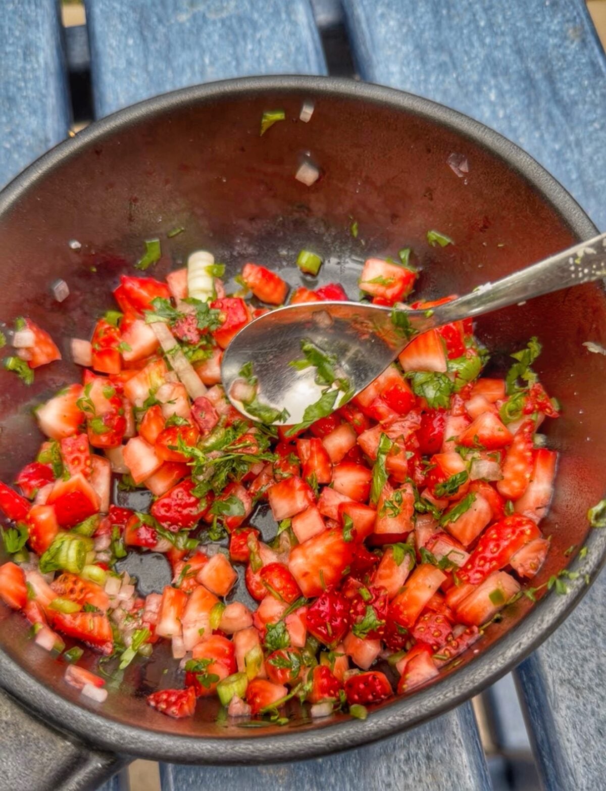 Fresh strawberry salsa with jalapeño, cilantro, onion, and green onions mixed together in a dark bowl with a spoon.