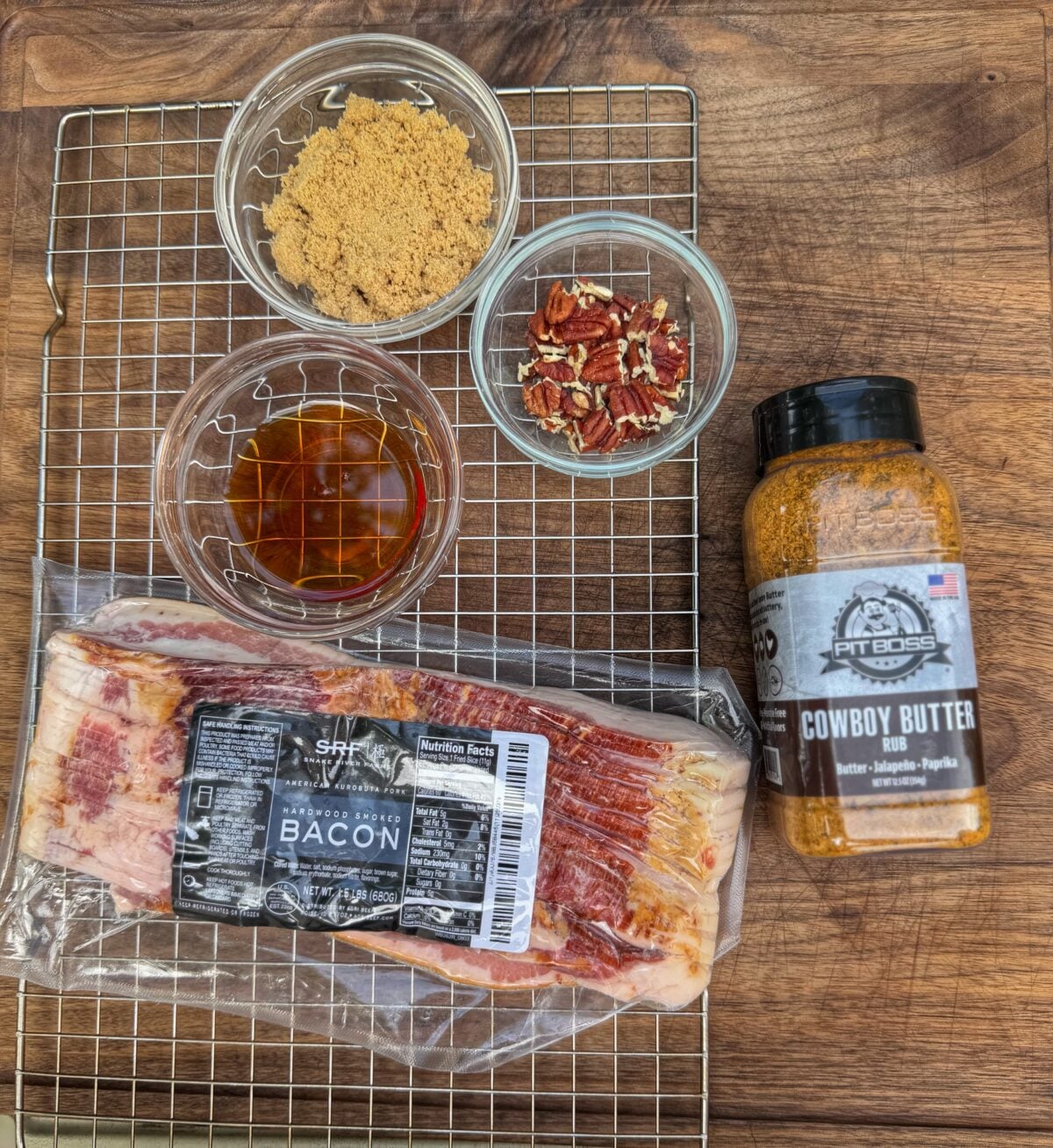 Ingredients for candied bacon including Snake River Farms bacon, brown sugar, maple syrup, chopped pecans, and Pit Boss Cowboy Butter seasoning on a wire rack over a wooden cutting board.
