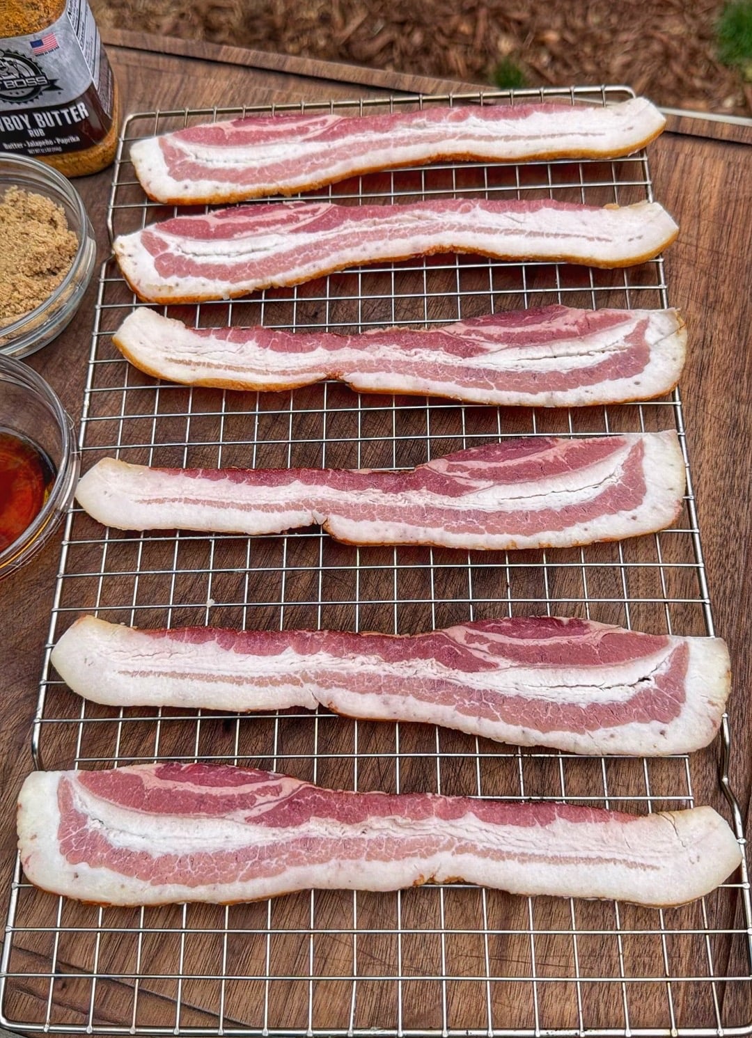 Raw thick cut bacon strips arranged on a wire rack over a wooden board with bowls of brown sugar and maple syrup nearby.