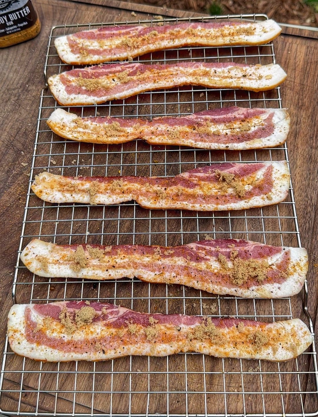 Raw bacon strips topped with brown sugar and seasoning on a wire rack over a wooden cutting board.