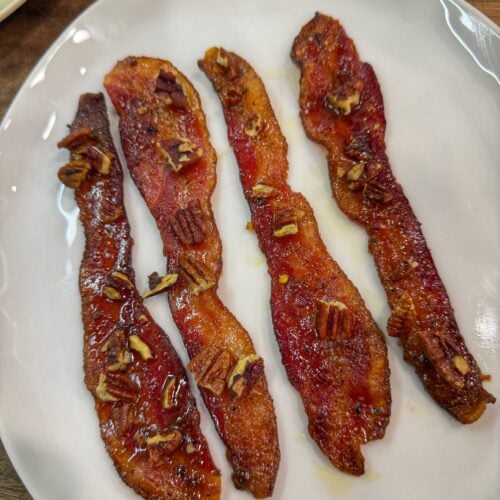 Cooked candied bacon strips topped with chopped pecans on a white plate.