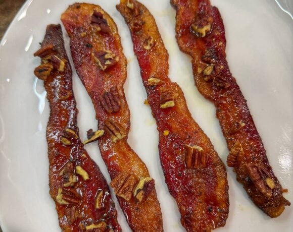 Cooked candied bacon strips topped with chopped pecans on a white plate.