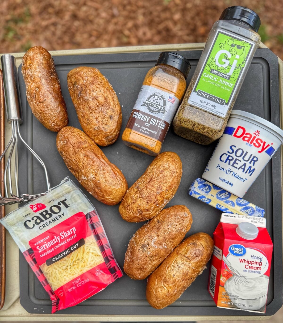 Ingredients for twice baked potatoes including russet potatoes, shredded cheddar cheese, sour cream, butter, heavy cream, Cowboy Butter seasoning, Garlic Junkie seasoning, and a potato masher on a tray.