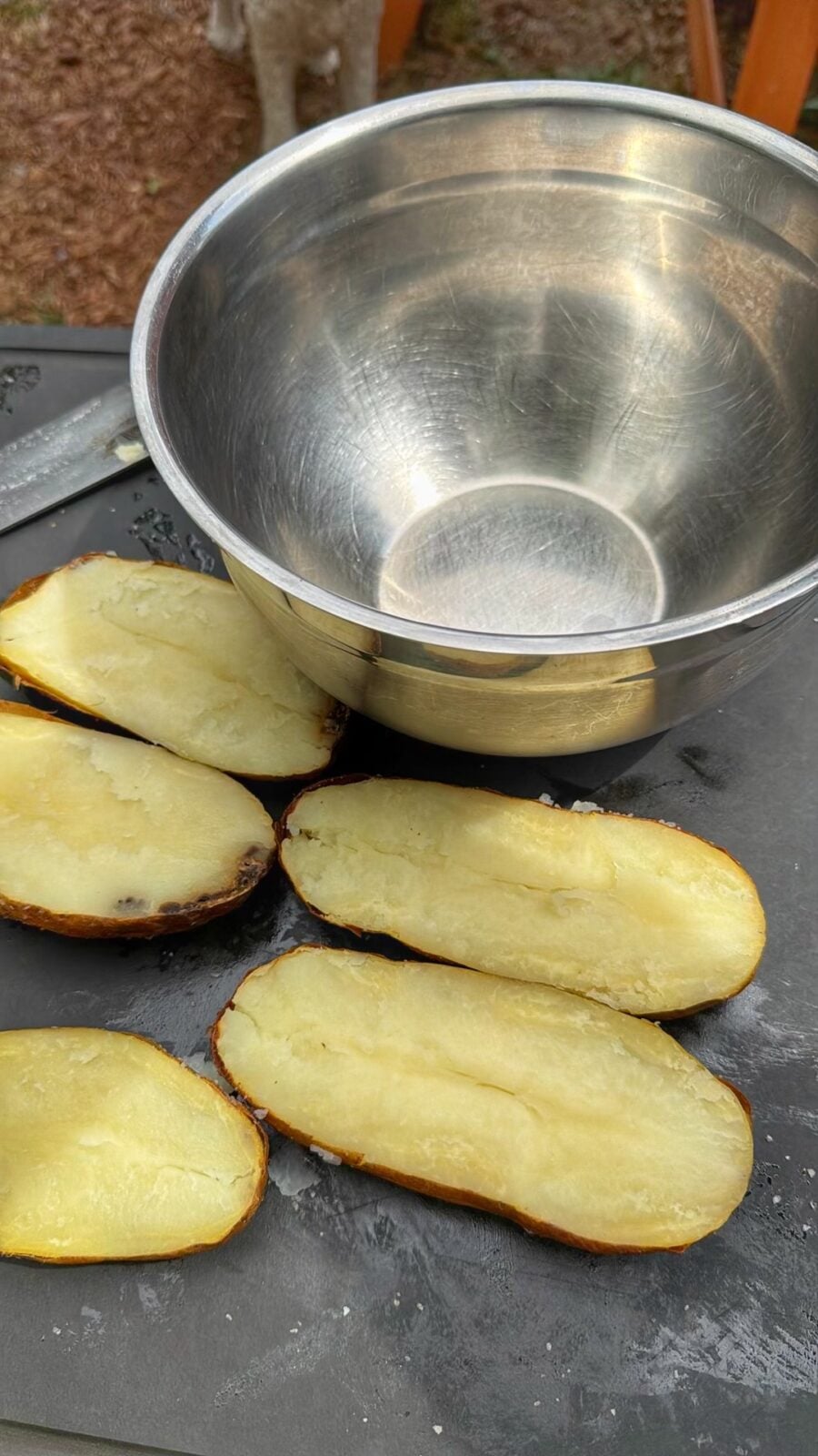 Baked russet potatoes cut in half next to a large stainless steel mixing bowl on an outdoor prep station.