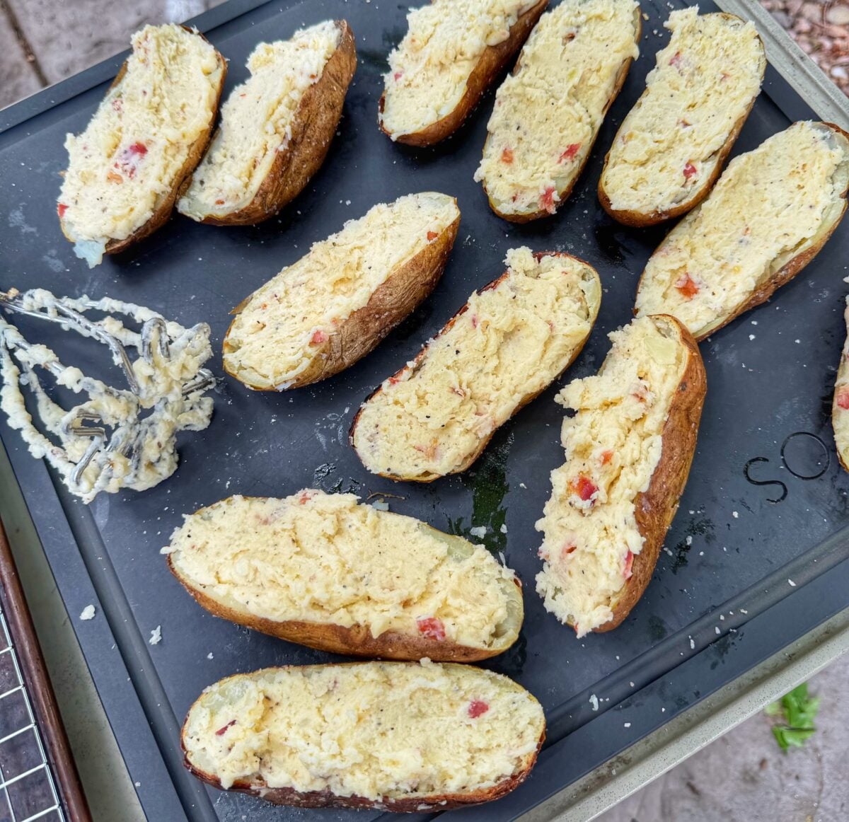 Potato skins filled with mashed potato mixture arranged on a prep tray beside a potato masher.