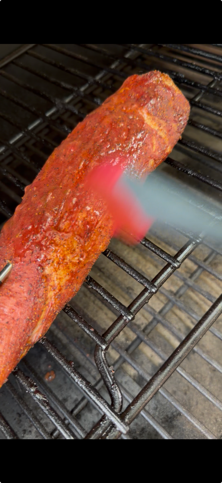 Strawberry glaze being brushed onto a seasoned pork tenderloin on the grill grates of a pellet smoker.