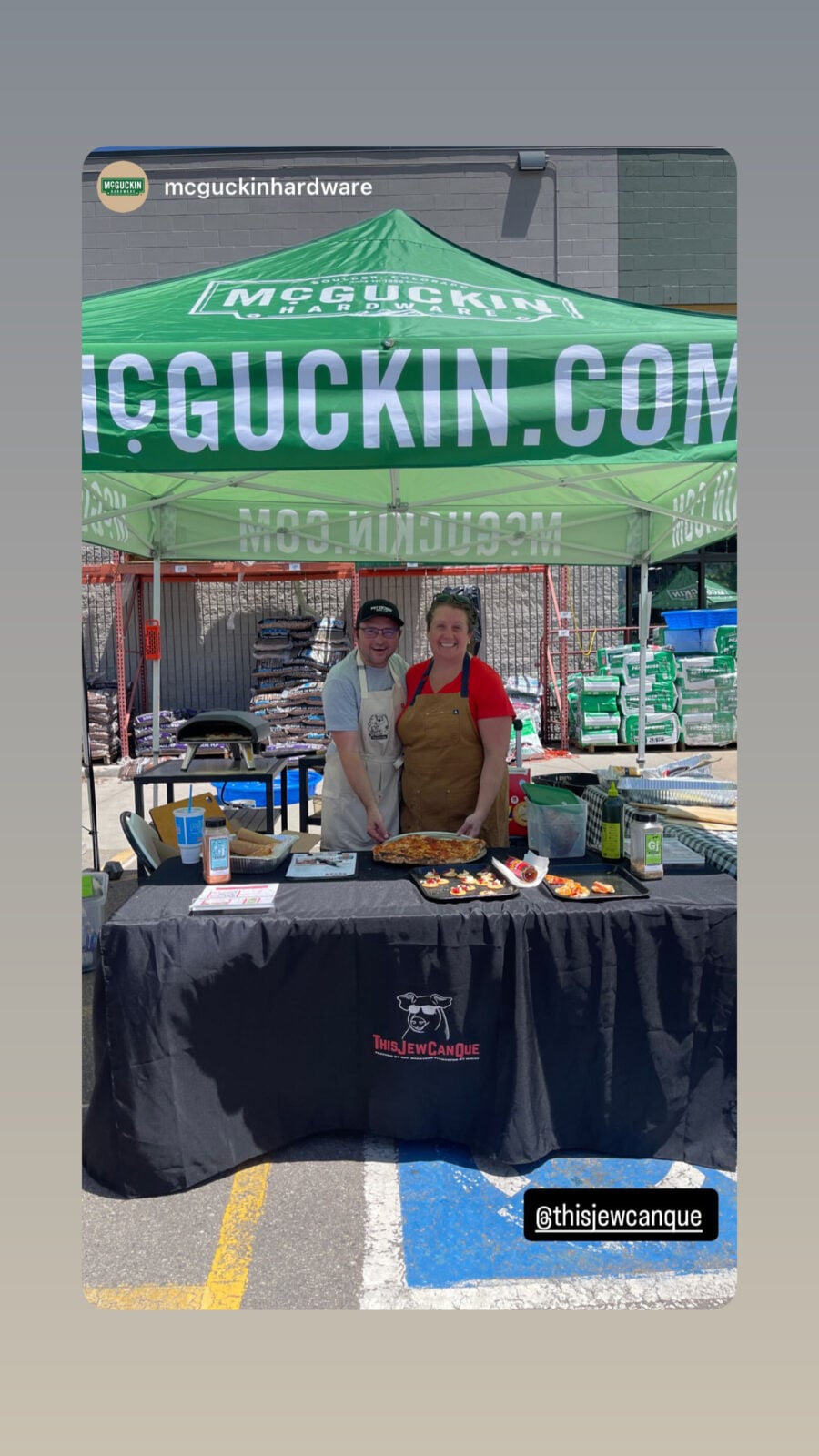 Adam McKenzie and his wife Isabel standing behind the ThisJewCanQue booth under a McGuckin Hardware tent with pizza samples and Ooni ovens.