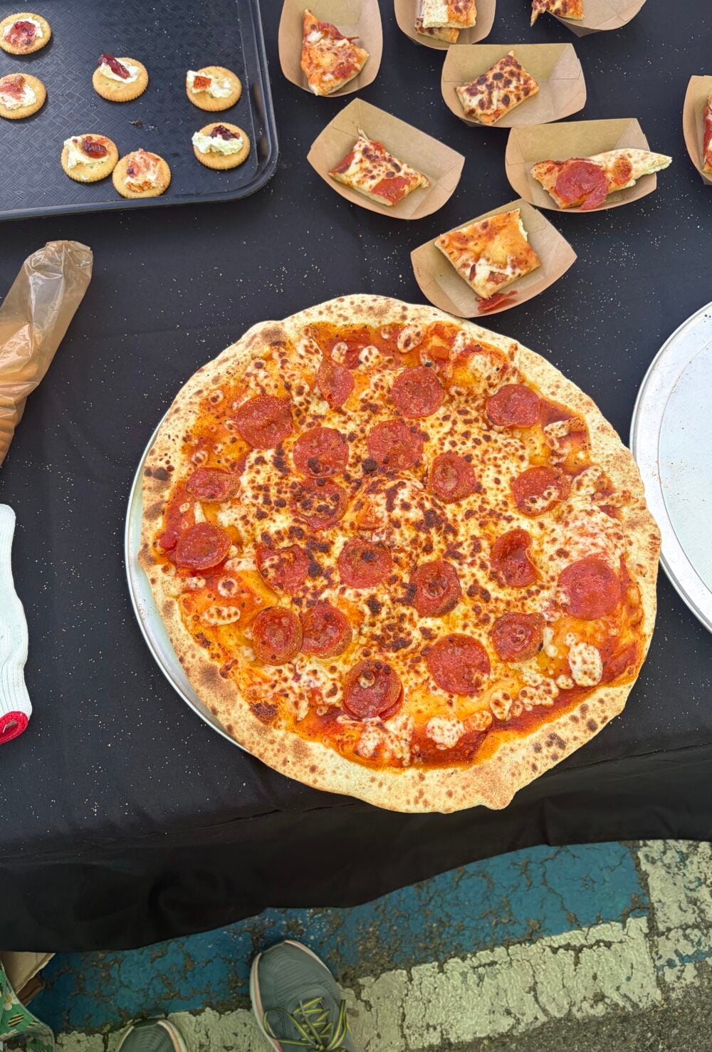Whole pepperoni pizza on a serving tray surrounded by sample slices and smoked cream cheese crackers.