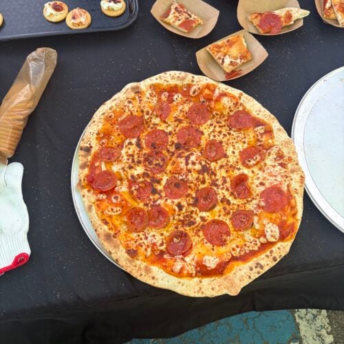 Whole pepperoni pizza on a serving tray surrounded by sample slices and smoked cream cheese crackers.