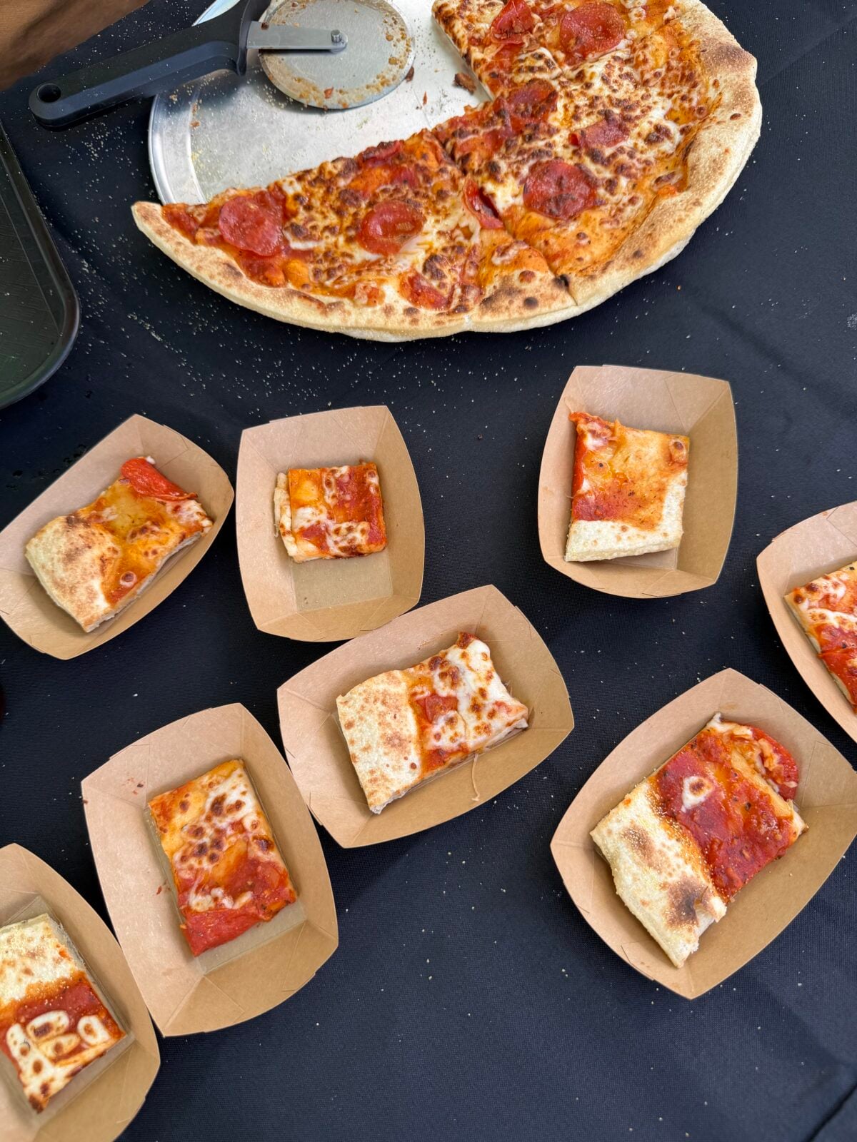 Sample-sized slices of pepperoni pizza served in brown paper food boats on a black tablecloth.