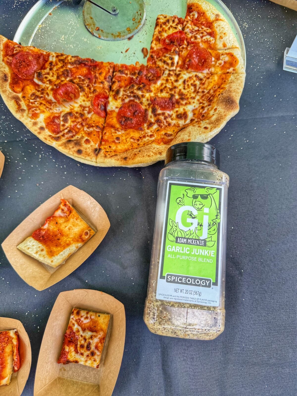 Container of Garlic Junkie seasoning next to pizza samples and a sliced pepperoni pizza.