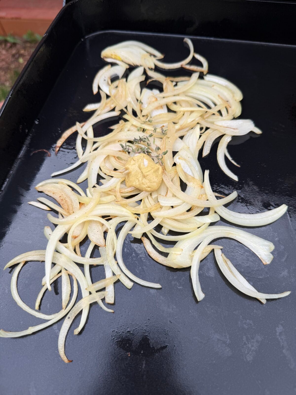 Sweet onions cooking on a Pit Boss griddle with Dijon mustard and fresh thyme for a French onion-inspired steak melt.
