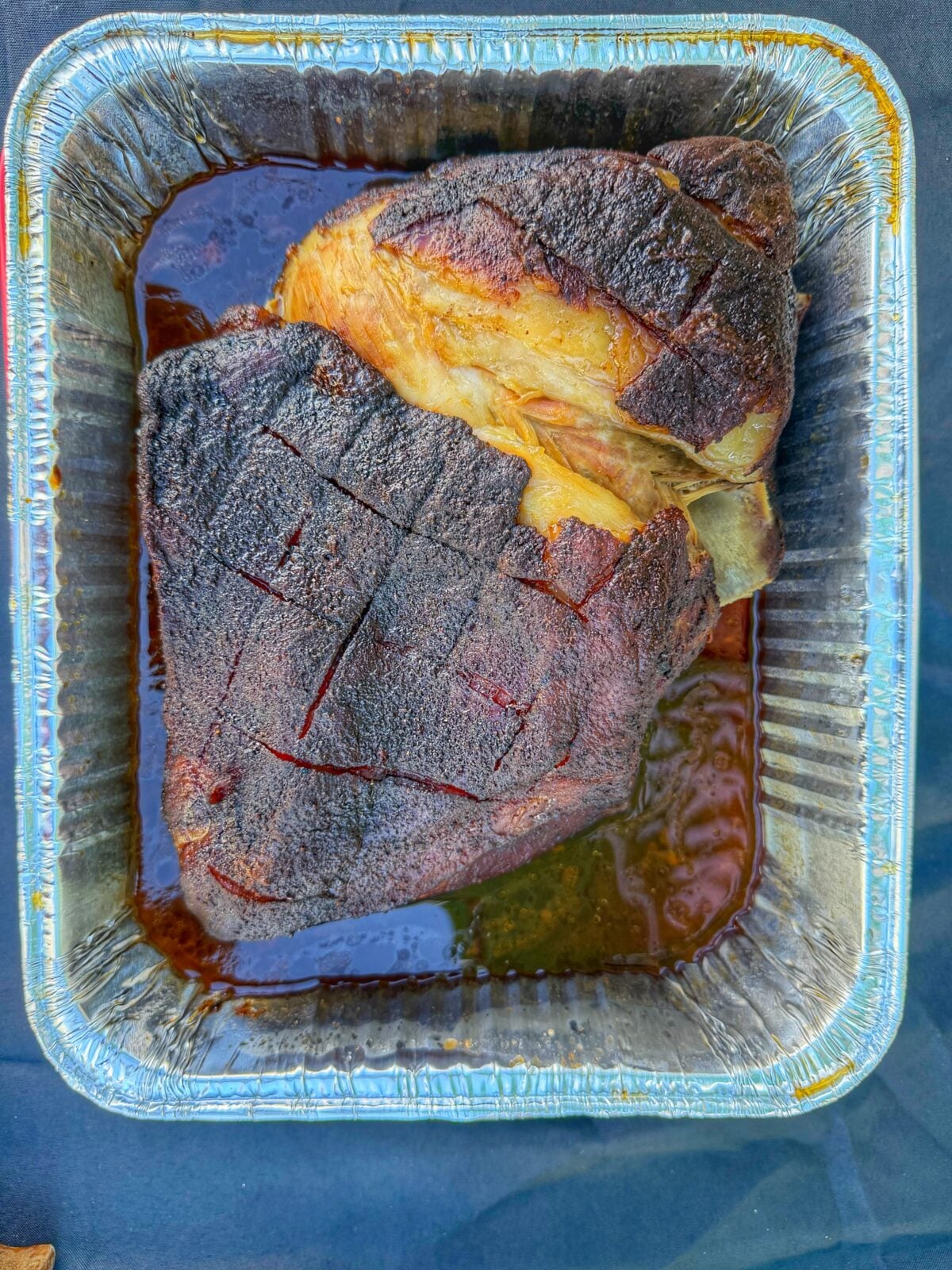 Two smoked pork shoulders resting in a foil pan with dark seasoned bark before being pulled.