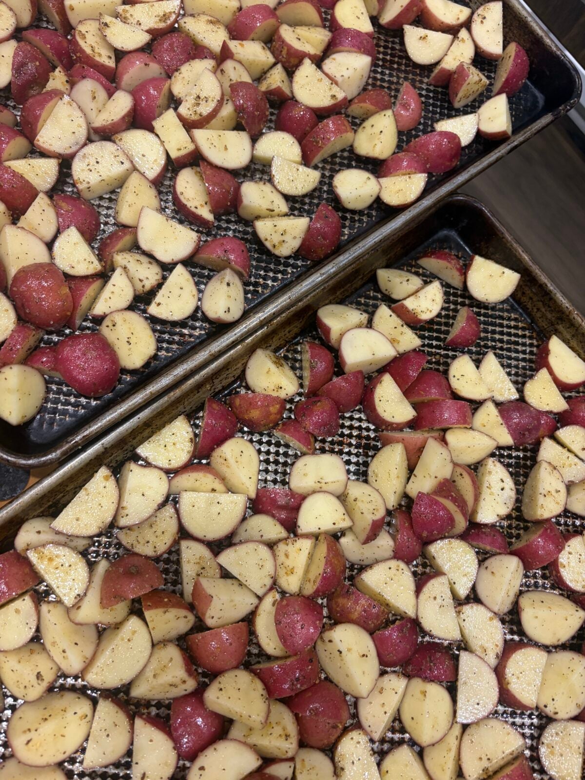 Seasoned quartered red potatoes spread across sheet pans and ready to cook.