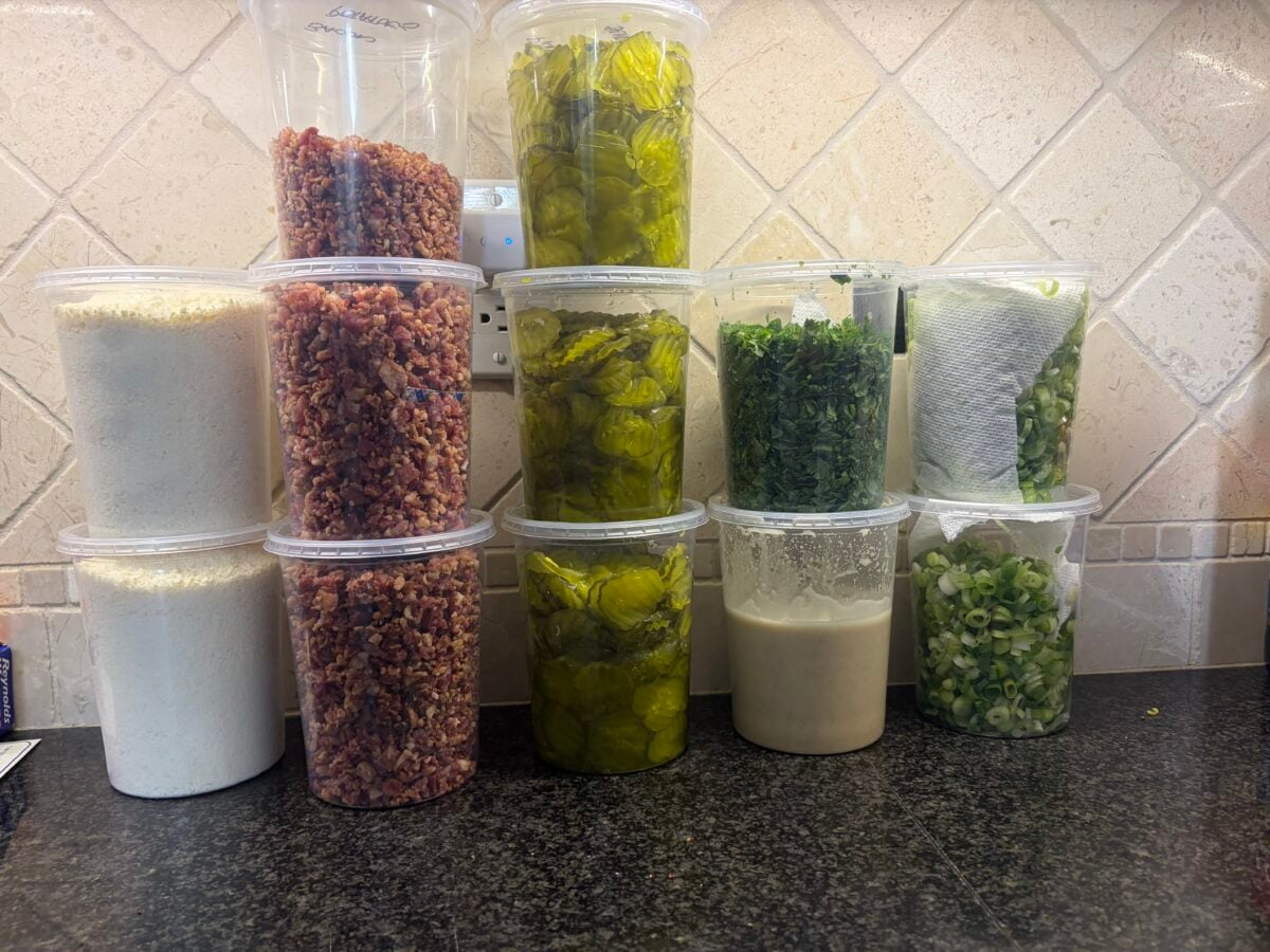 Containers of bacon, pickles, herbs, green onions and other toppings prepped for a large outdoor cooking demo.